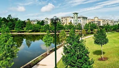 Walking path beside a pond with trees and mid-rise buildings in The Woodlands, Texas